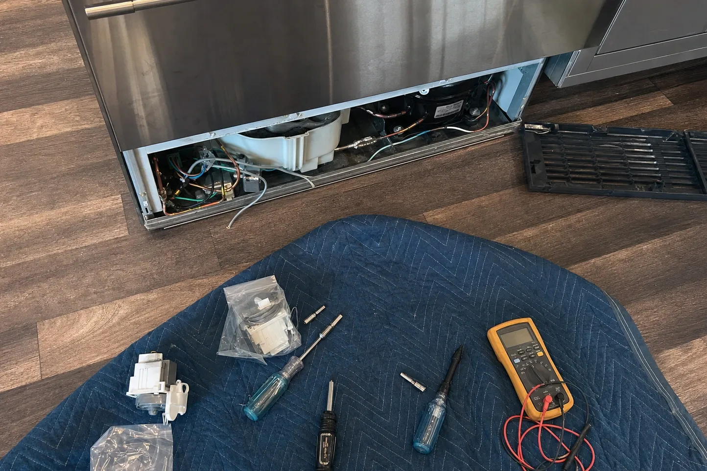 Lower grille removed from a tall stainless built-in, exposing the compressor and condenser fan, with a multimeter, drivers and bagged parts on a blanket alongside