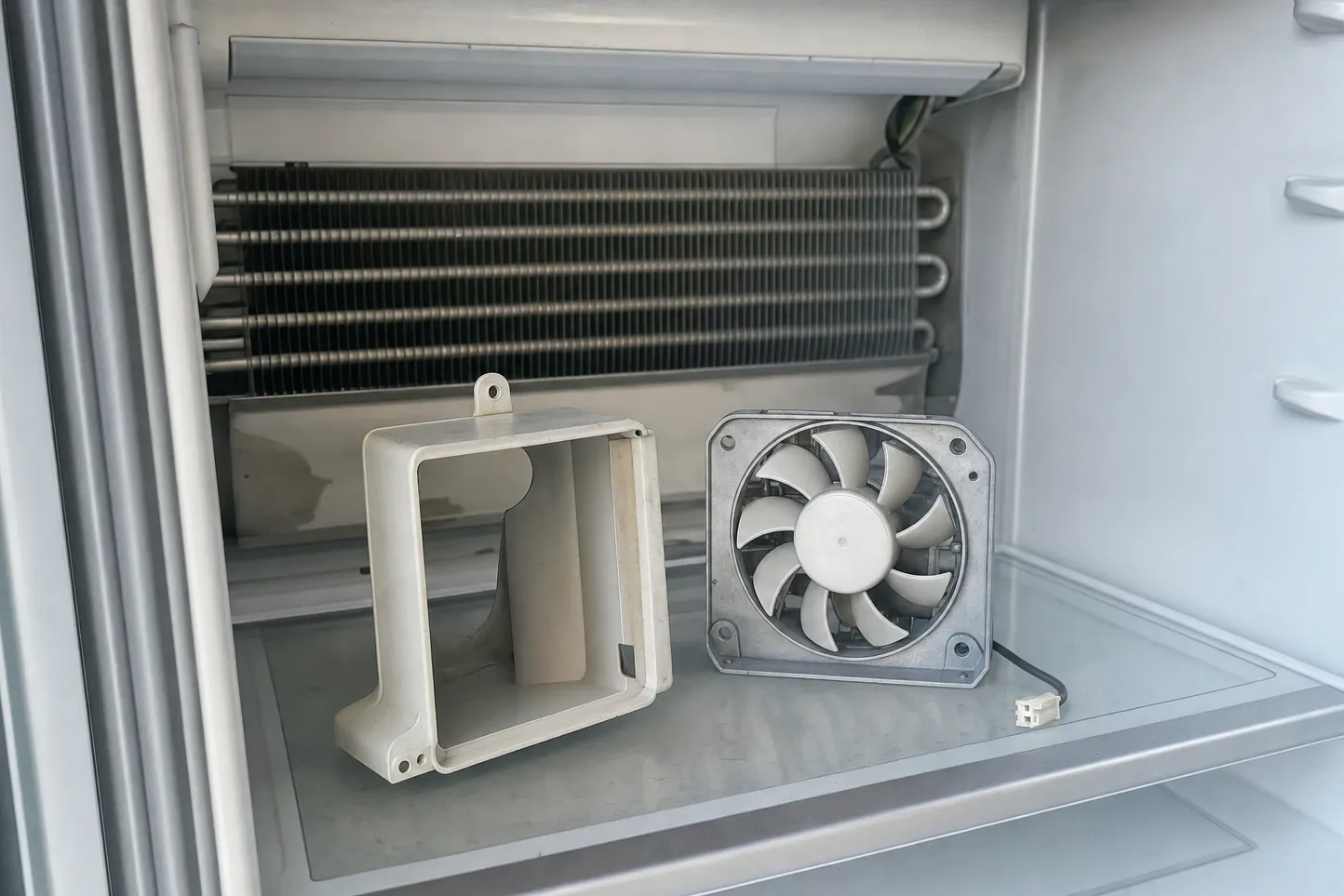 Finned evaporator coil exposed across the rear wall of a white compartment, with the plastic fan shroud and the axial fan lifted out and resting on the shelf below