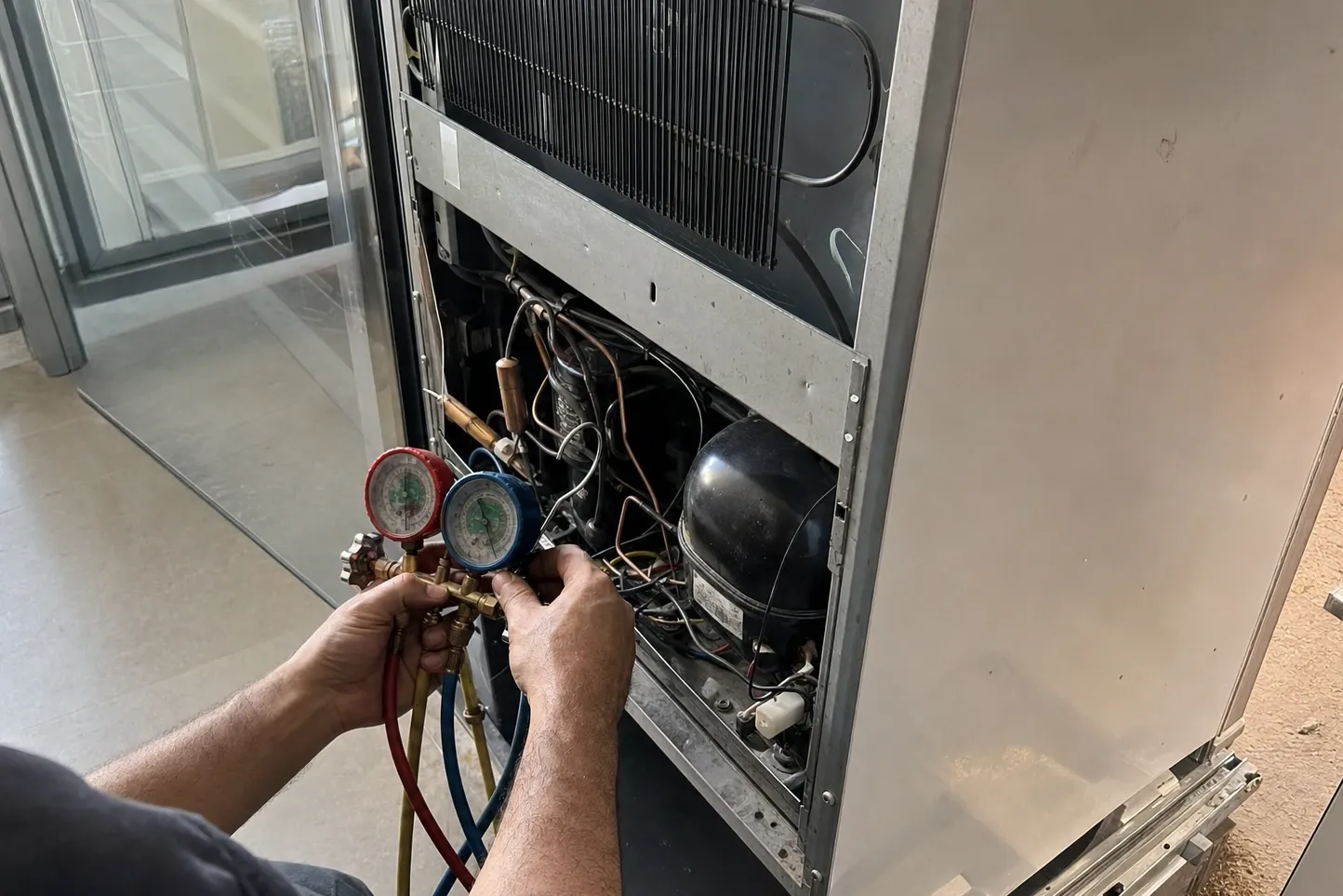 Hands holding a red and blue manifold gauge set at the opened rear machine compartment of a tall stainless cabinet, hoses running down to the black compressor below the condenser coil